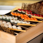 Plateau en bois présentant un assortiment de sushis, comprenant des nigiris de saumon, de thon et de crevette, ainsi que des makis coupés en morceaux, disposés en rangées avec des baguettes posées à côté.
