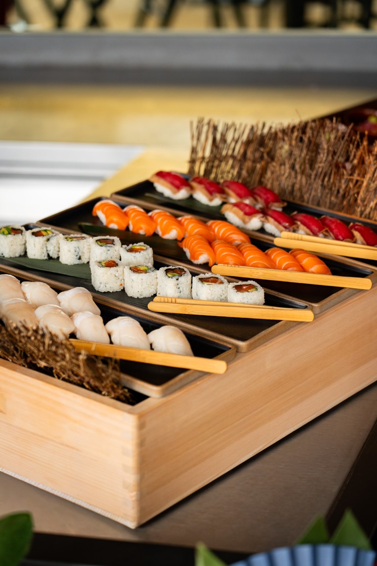 Plateau en bois présentant un assortiment de sushis, comprenant des nigiris de saumon, de thon et de crevette, ainsi que des makis coupés en morceaux, disposés en rangées avec des baguettes posées à côté.