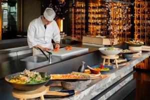 Chef en tenue blanche découpant du saumon sur un comptoir de cuisine ouverte, entouré de plats élégamment dressés, avec un décor moderne et chaleureux comprenant des étagères éclairées et des branches fleuries.