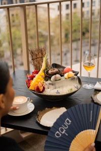 Une personne est assise à une table en extérieur, tenant une tasse de café, tandis qu’un élégant plateau de desserts et de fruits est présenté sur glace au centre de la table. À côté, un verre de vin blanc et un éventail reposent, avec en arrière-plan une vue urbaine floue depuis un balcon.